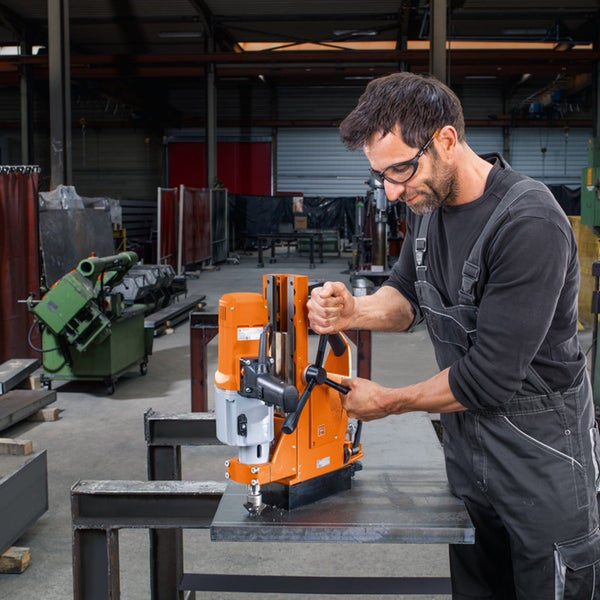 Ein Mann mit Schutzbrille und Arbeitslatzhose bedient eine orangefarbene Magnet-Kernbohrmaschine auf einer Stahlplatte in einer Werkstatt.