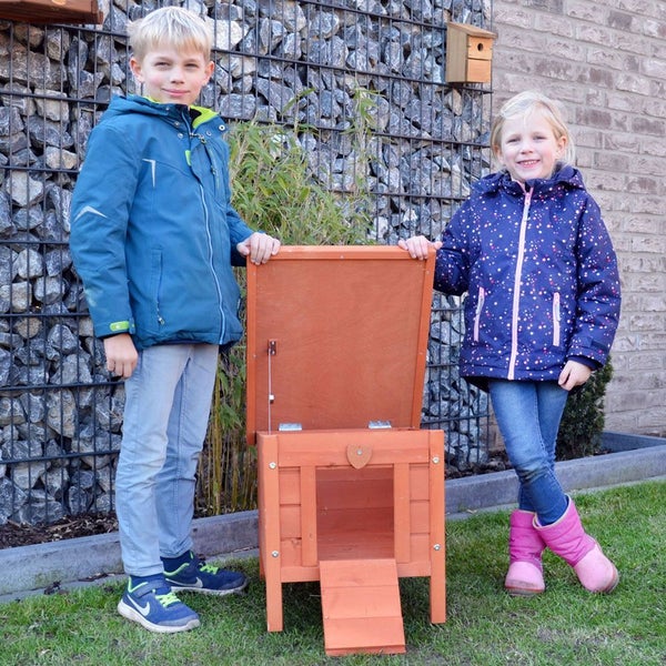 Kleintierhaus aus Holz mit aufklappbarem Dach und zwei Kindern in einem Garten vor einer Steinkorbwand.