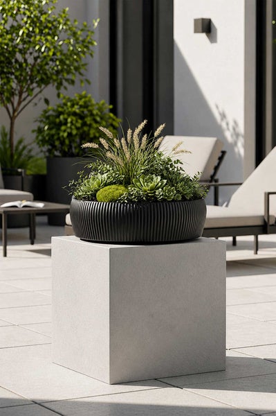 Schwarze Pflanzschale mit Rillenstruktur, bepflanzt mit Sukkulenten und Ziergräsern, auf einem grauen Betonsockel auf einer modernen Terrasse.