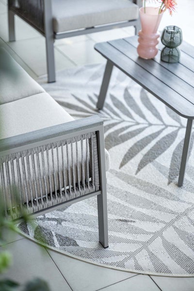 Modernes Outdoor-Loungeset mit grauen Seilmöbeln, hellen Polstern, einem Couchtisch und einem runden Teppich mit Palmenblattmuster.