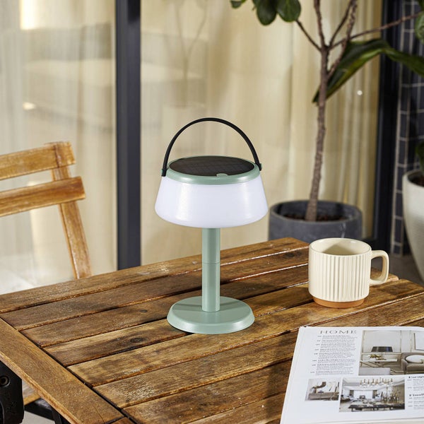 Grüne Solar-LED-Tischleuchte mit Tragegriff auf einem Holztisch im Außenbereich neben einer Tasse und einer Zeitschrift.