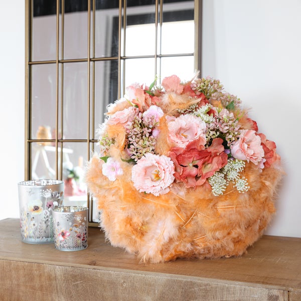 Dekoratives Gesteck aus rosa Kunstblumen in einer Basis aus orangefarbenen Federn, neben zwei gemusterten Teelichthaltern aus Glas vor einem Spiegel.