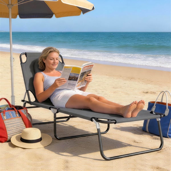 Eine Frau liegt auf einer grauen, klappbaren Sonnenliege am Strand unter einem Sonnenschirm und liest.