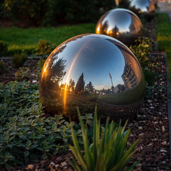 Drei reflektierende Gartenkugeln aus Edelstahl in einem hellen, organisierten Gartenbeet mit Pflanzen und Mulch.