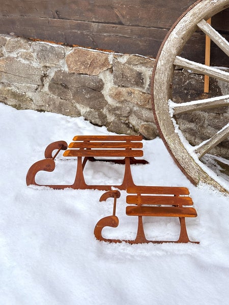 Zwei dekorative Schlitten aus Metall mit Edelrostpatina im Schnee vor einer Steinmauer mit einem alten Wagenrad.