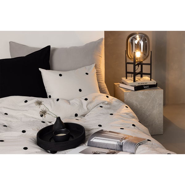 Schlafzimmer mit gepunkteter Bettwäsche, Kissen, schwarzem Tablett auf dem Bett und Glas-Tischleuchte auf einem Beistelltisch aus Stein.