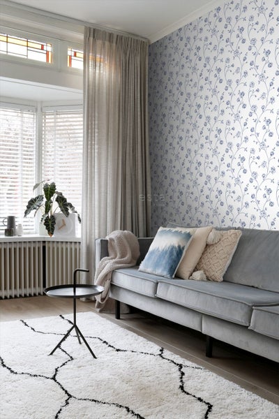 Vliestapete mit blauem Blumenmuster in einem hellen, modern eingerichteten Wohnzimmer mit grauem Sofa und weißem Teppich.