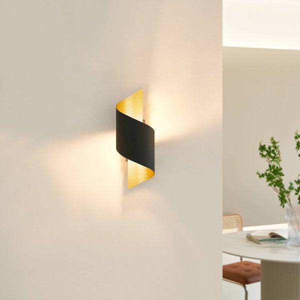 Moderne Wandleuchte mit geschwungenem Gehäuse in Schwarz und Gold, montiert an einer hellen Wand, strahlt warmes Licht aus.