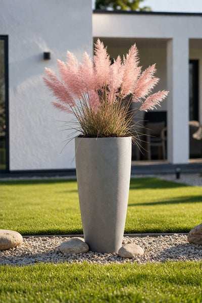 Hoher runder Pflanzkübel in Betonoptik mit rosa Pampasgras auf einer Kiesfläche in einem modernen Garten.