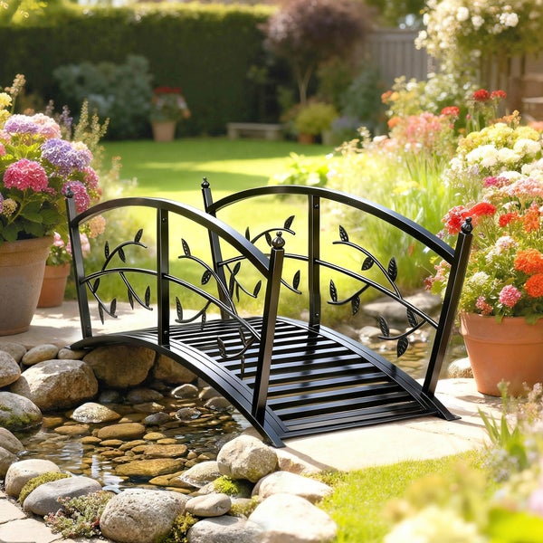 Schwarze Gartenbrücke aus Metall mit Blattornamenten über einem kleinen Gartenteich in einer hellen Gartenlandschaft.