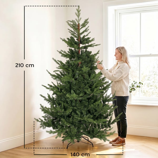 Eine Frau richtet einen 210 Zentimeter hohen und 140 Zentimeter breiten künstlichen Weihnachtsbaum in einem hellen Wohnraum aus.