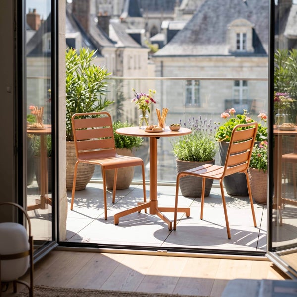 Moderner, heller Balkon mit zwei Metallstühlen, einem runden Tisch und verschiedenen Topfpflanzen vor einer Stadtkulisse.