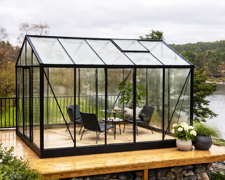Großes Gewächshaus mit schwarzem Aluminiumrahmen und Glaswänden, als Sitzbereich auf einer Holzterrasse am See eingerichtet.