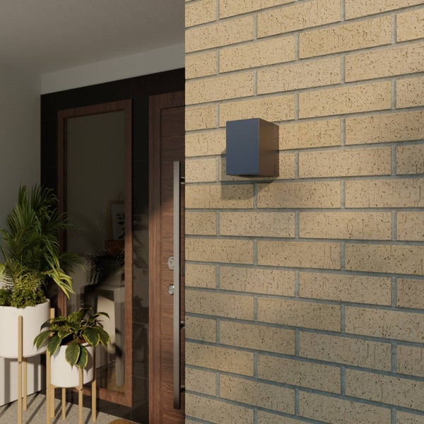 Moderne würfelförmige Außenwandleuchte montiert an einer Ziegelwand neben einer Eingangstür mit Pflanzen.