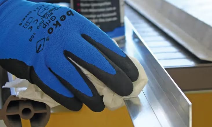 Eine Hand in einem blau-schwarzen beko Grip Arbeitshandschuh reinigt ein silbernes Metallprofil mit einem weißen Tuch.
