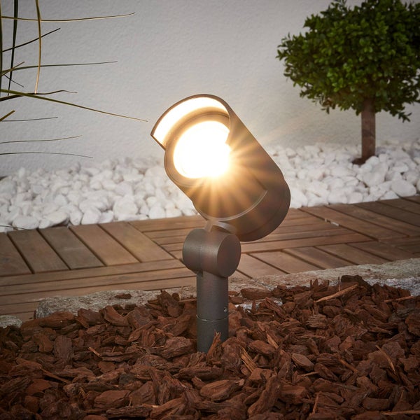 Moderner LED-Gartenstrahler aus Metall mit Erdspieß, installiert in Rindenmulch neben einer Holzterrasse und weißen Kieseln.