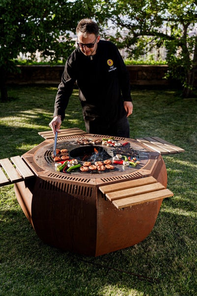Ein Koch bereitet auf einem runden Feuerschalengrill mit Holzablagen im Garten Speisen zu.