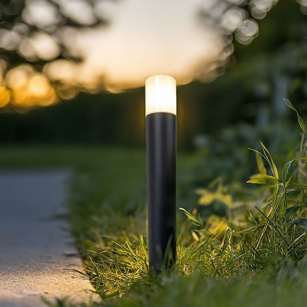 Schwarze zylindrische Außenleuchte aus Metall, beleuchtet in einem Garten an einem Weg.