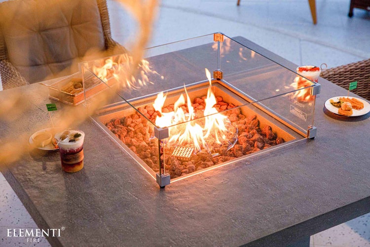 Gaskamin-Tisch in Steinoptik mit Glas-Windschutz, Lavasteinen und hellem Feuer auf einer Terrasse. Elementi Fire Logo.