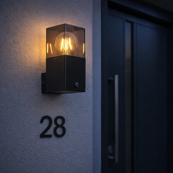 Moderne Außenwandleuchte mit Sensor, schwarzem Sockel und Glasgehäuse an einer grauen Fassade über der Hausnummer achtundzwanzig.