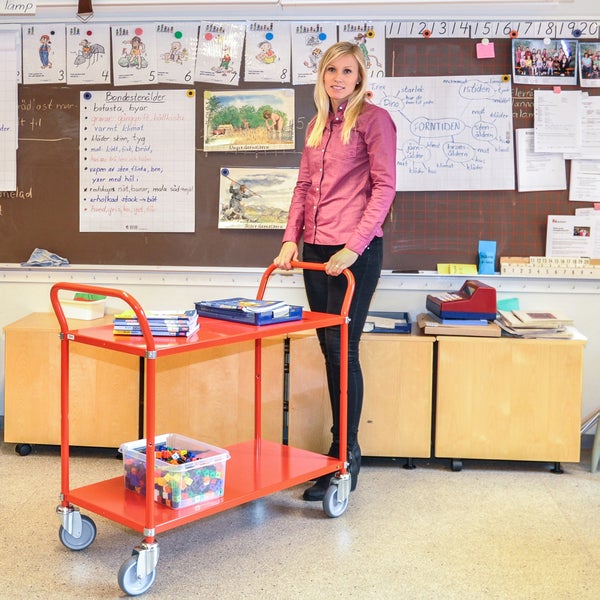 Szene eines Klassenzimmers mit einer Lehrerin und einem Servierwagen mit zwei Ablagen, beladen mit Büchern und Spielzeug.