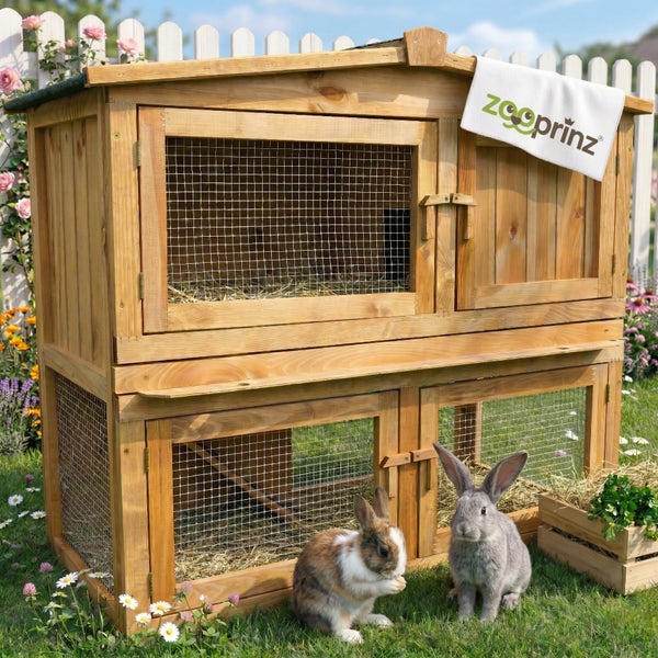 Zweistöckiger Kleintierstall aus Holz mit Drahtgitter im Garten, zwei Kaninchen davor auf der Wiese, Zooprinz Logo.