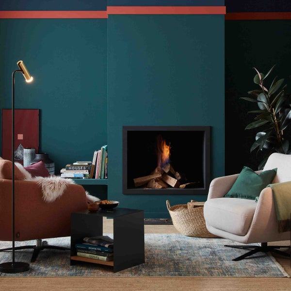 Modernes Wohnzimmer mit Kamin, brennendem Holz, zwei Sesseln, Beistelltisch, Stehlampe und dunklen Wänden mit terrakottafarbenem Akzentstreifen.