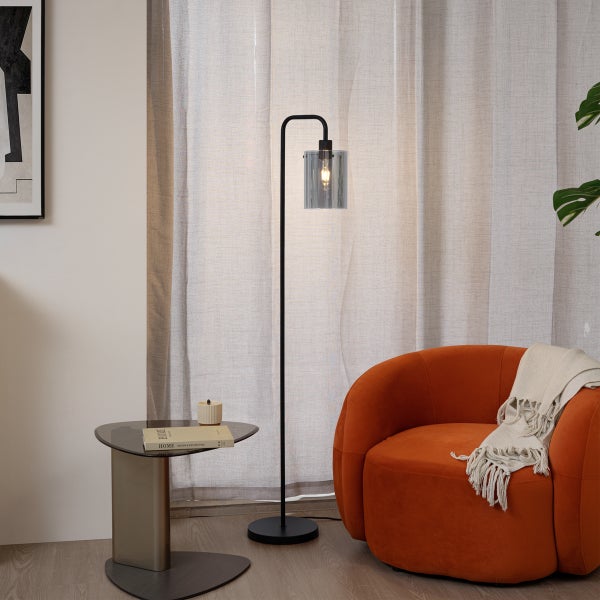 Moderne Stehleuchte mit zylindrischem Glasschirm und schwarzem Metallgestell in einem Wohnzimmer neben einem orangefarbenen Sessel.