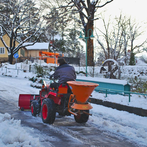 Ein Person räumt mit einem Traktor mit Schneeschild und Streuaufsatz einen verschneiten Weg im Winter.