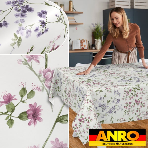 Abwaschbare Tischdecke mit rosa und lila Blumen. Eine Frau legt die Decke auf einen Holztisch in einer hellen Küche. ANRO Deutsche Manufaktur Logo.