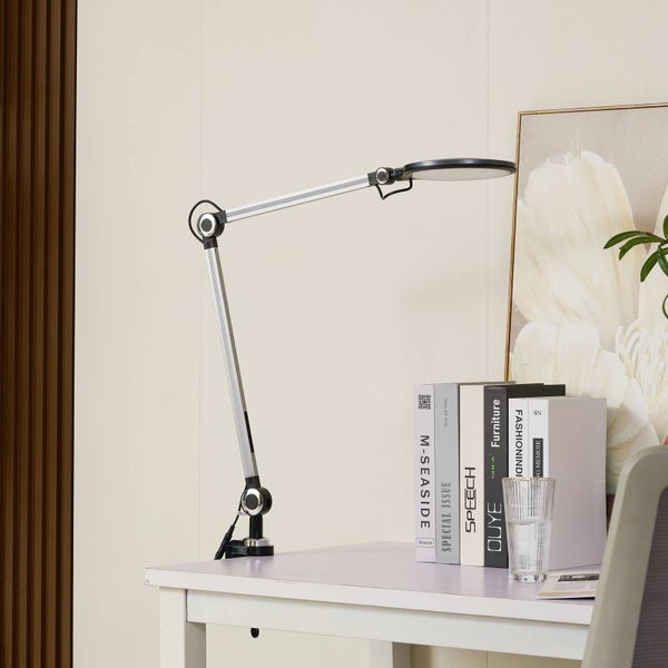 Verstellbare Schreibtischlampe mit silbernem Gelenkarm und schwarzem, rundem Lampenkopf, montiert an einem weißen Tisch neben Büchern.