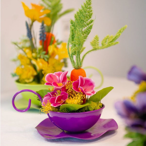 Ostergesteck in einer metallenen Tasse als Pflanzgefäß mit Kunstblumen, Moos und einer Möhre als Frühlingsdekoration für den Tisch.