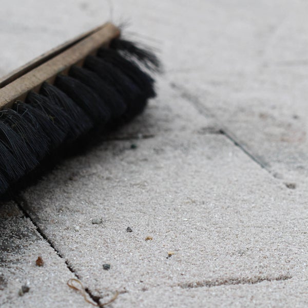 Ein Holzbesen mit schwarzen Borsten fegt feinen Fugensand in die Zwischenräume von Gehwegplatten.
