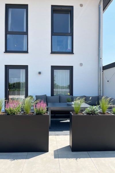 Moderne Terrasse mit großen schwarzen Pflanzkübeln, Gräsern, Heidekraut, dunkelgrauem Lounge-Sofa und Tisch vor einer Hauswand.