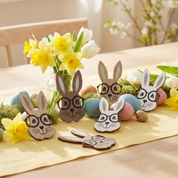 Osterdekoration mit hölzernen Osterhasenfiguren mit Brille, Eiern und einer Vase mit Narzissen auf einem Tisch.