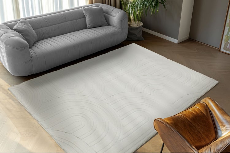 Ein moderner Wohnzimmerteppich mit geometrischem Reliefmuster auf einem Boden mit Fischgrätparkett vor einem Sofa.