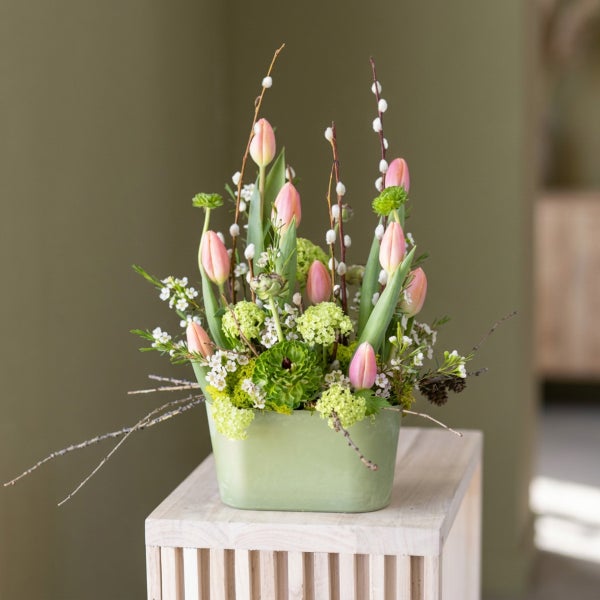 Frühlingshafter Blumenstrauß aus rosa Tulpen, grünen Blüten und Weidenkätzchen in einer rechteckigen Pflanzschale auf einem Holzsockel.