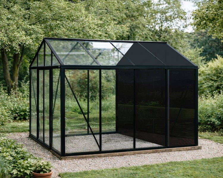 Gewächshaus mit schwarzem Metallrahmen und Glasscheiben, teilweise getönt, auf Kiesfläche in einem grünen Garten.