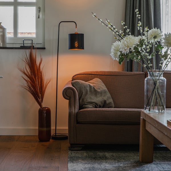 Modernes Wohnzimmer mit schwarzer Stehleuchte, braunem Sofa und Holzmöbeln. Warmes Licht und dekorative Vasen mit Blumen und Gräsern.