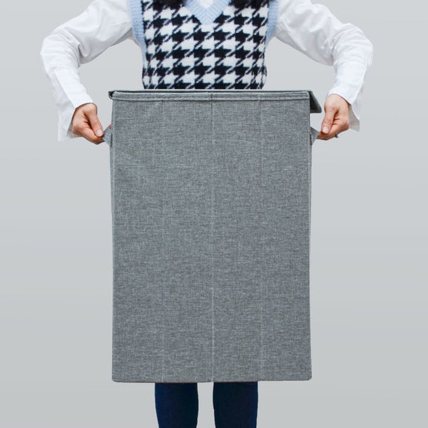 Grauer faltbarer Wäschekorb aus Stoff mit Deckel und seitlichen Griffen, von einer Person gehalten.