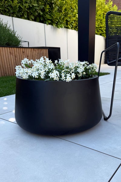 Runder schwarzer Pflanzkübel mit weißen Blumen auf einer modern gefliesten Terrasse im Garten.