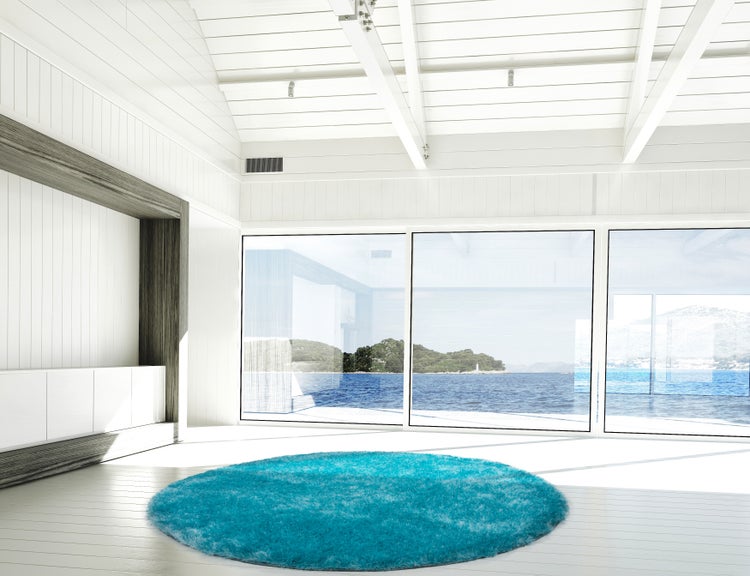 Heller moderner Wohnraum mit rundem türkisfarbenem Hochflorteppich und großen Fensterfronten mit Blick auf das Meer.