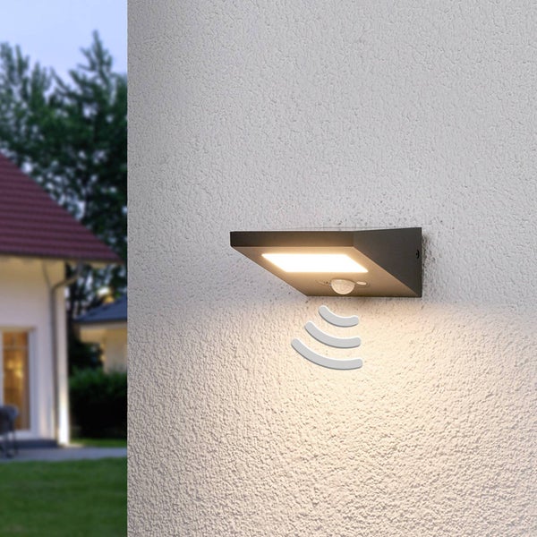 LED-Außenwandleuchte in Anthrazit mit Bewegungsmelder an einer weißen Hauswand, warmweißes Licht, Garten im Hintergrund bei Dämmerung.