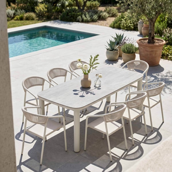 Helles Gartenmöbel-Set auf einer geräumigen Terrasse am Pool, bestehend aus modernem Tisch und acht Stühlen mit Seilbespannung und Metallgestell.