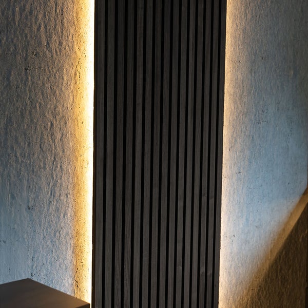 Akustikpaneel mit dunklen Holzlamellen und indirekter LED-Beleuchtung an einer Wand mit Struktur.