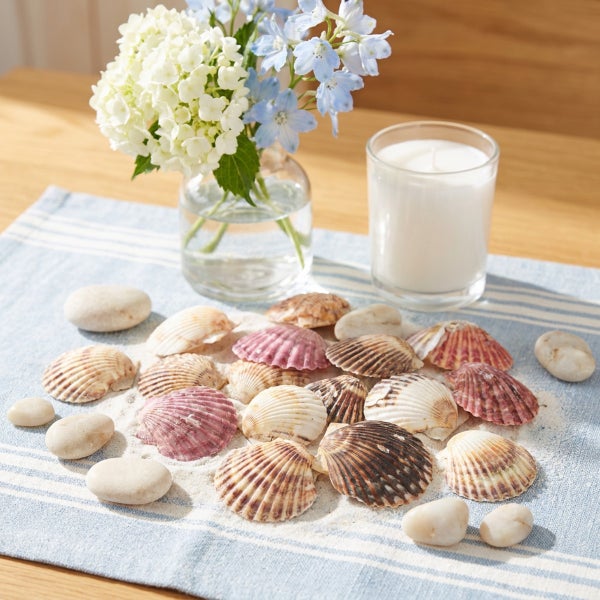 Dekorative Muscheln und Kieselsteine auf einer gestreiften Tischdecke, arrangiert mit einer Blumenvase und einer Kerze im Glas als Tischdeko.