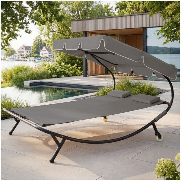 Moderne Doppel-Sonnenliege mit verstellbarem Sonnendach, zwei Kissen und Rollen auf einer Terrasse neben einem Pool.