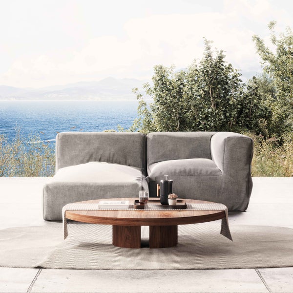 Moderne Outdoor-Lounge auf einer Terrasse mit Meerblick, grauem Modulsofa und rundem Holztisch auf einem gestreiften Teppich.