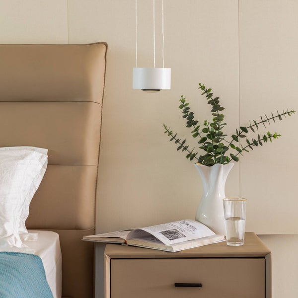 Modernes Schlafzimmer mit weißer Pendelleuchte über einem beigen Nachttisch mit Vase, Eukalyptuszweigen, Buch und Wasserglas neben dem Bett.