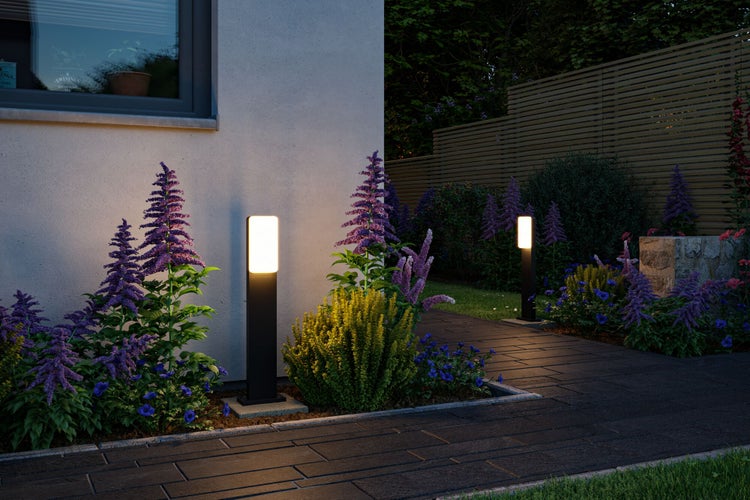 Moderne schwarze Wegeleuchten beleuchten einen gepflasterten Gartenweg bei Dämmerung neben Blumenbeeten und einer Hauswand.
